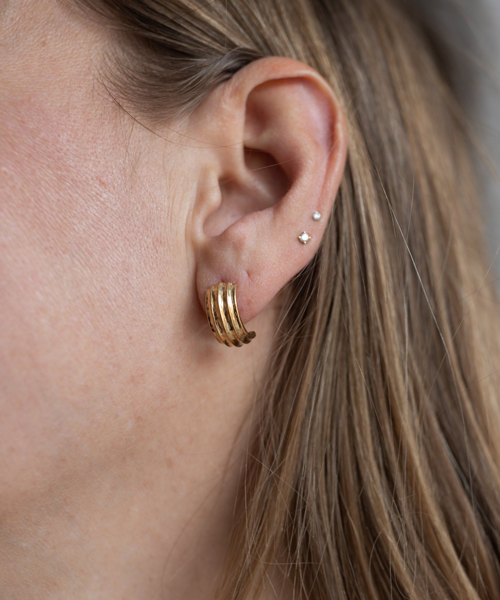 Close-up of a person’s ear with long blonde hair, wearing WALD Berlin's Pop Wave Earrings Small—an 18k gold plated creole hoop in the lobe and two small 925 silver pin studs in the upper ear.