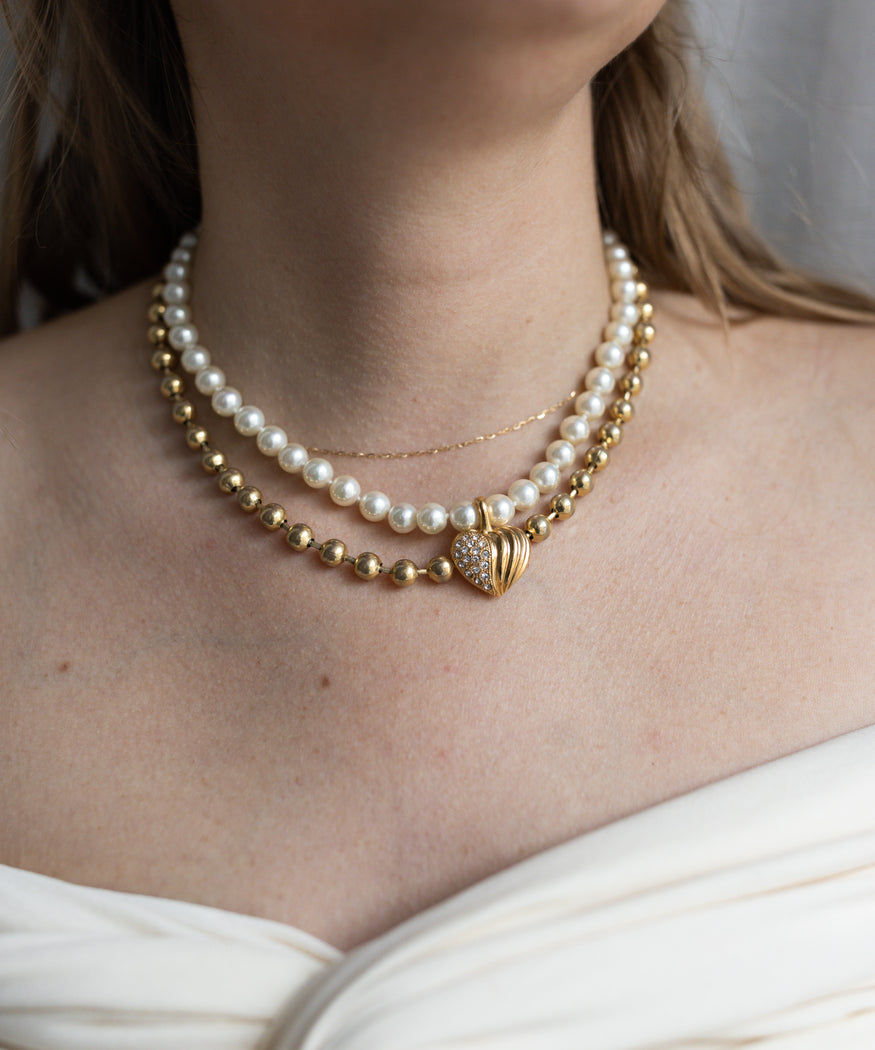 A woman wears the WALD Berlin Mom Wax Pearl Heart Necklace—featuring layered pearls, a gold-plated heart, and a fine gold chain—styled with an off-white, off-shoulder top.