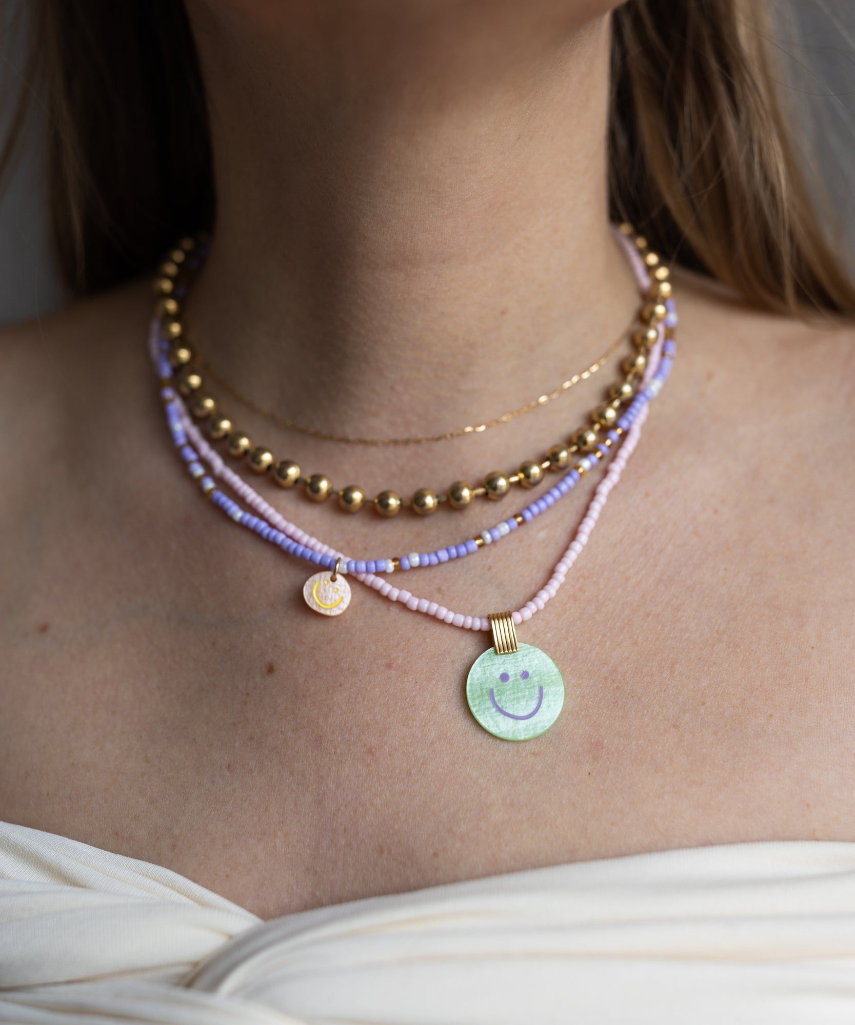 A woman layers WALD Berlin necklaces, including 18k gold plated beads, pastel beaded chains, and two round pendants—a Pop Smilie Mini medaillon and a classic smilie necklace—each featuring playful smiley face designs.