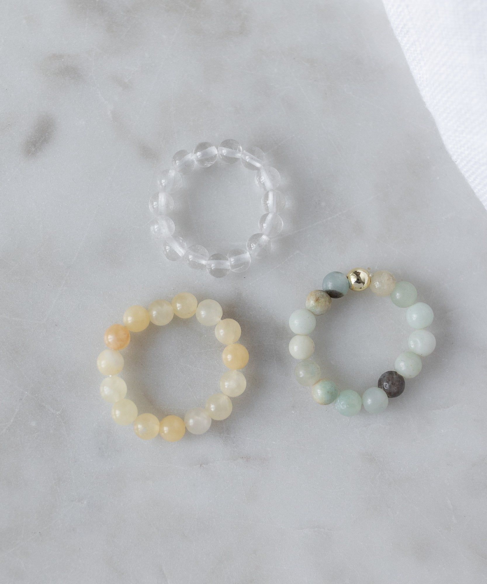 The WALD Berlin Pop Gemstone Ring Set features three beaded rings—a clear gemstone, a yellow band, and a multicolored ring with a gold bead—arranged on light marble. Their flexible sizing makes them charming gift ideas.
