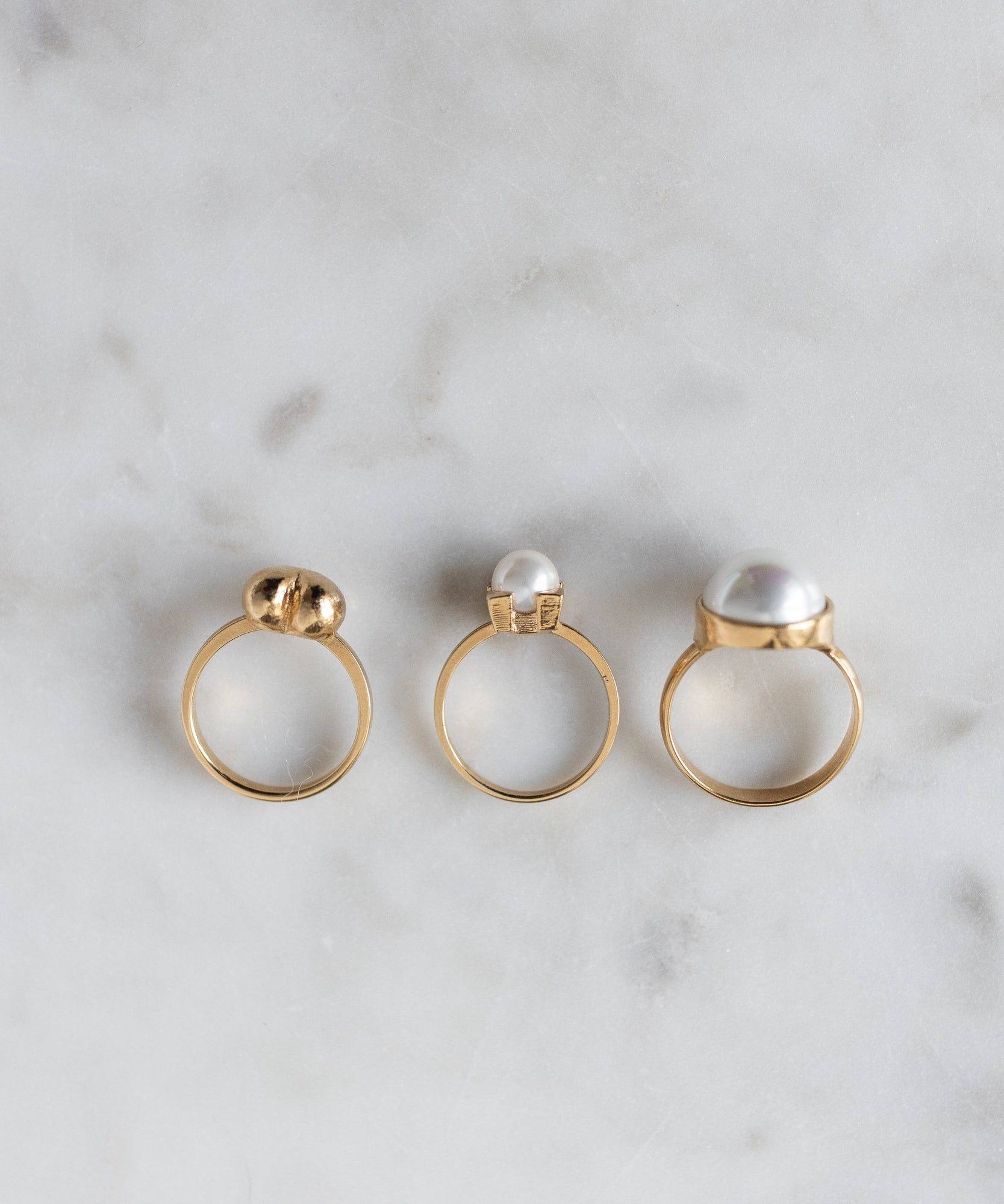 Three WALD Berlin Pop Pearl Ring Minis, crafted from 18k gold plated brass, are displayed in a row on light marble, creating a modern yet elegant arrangement.