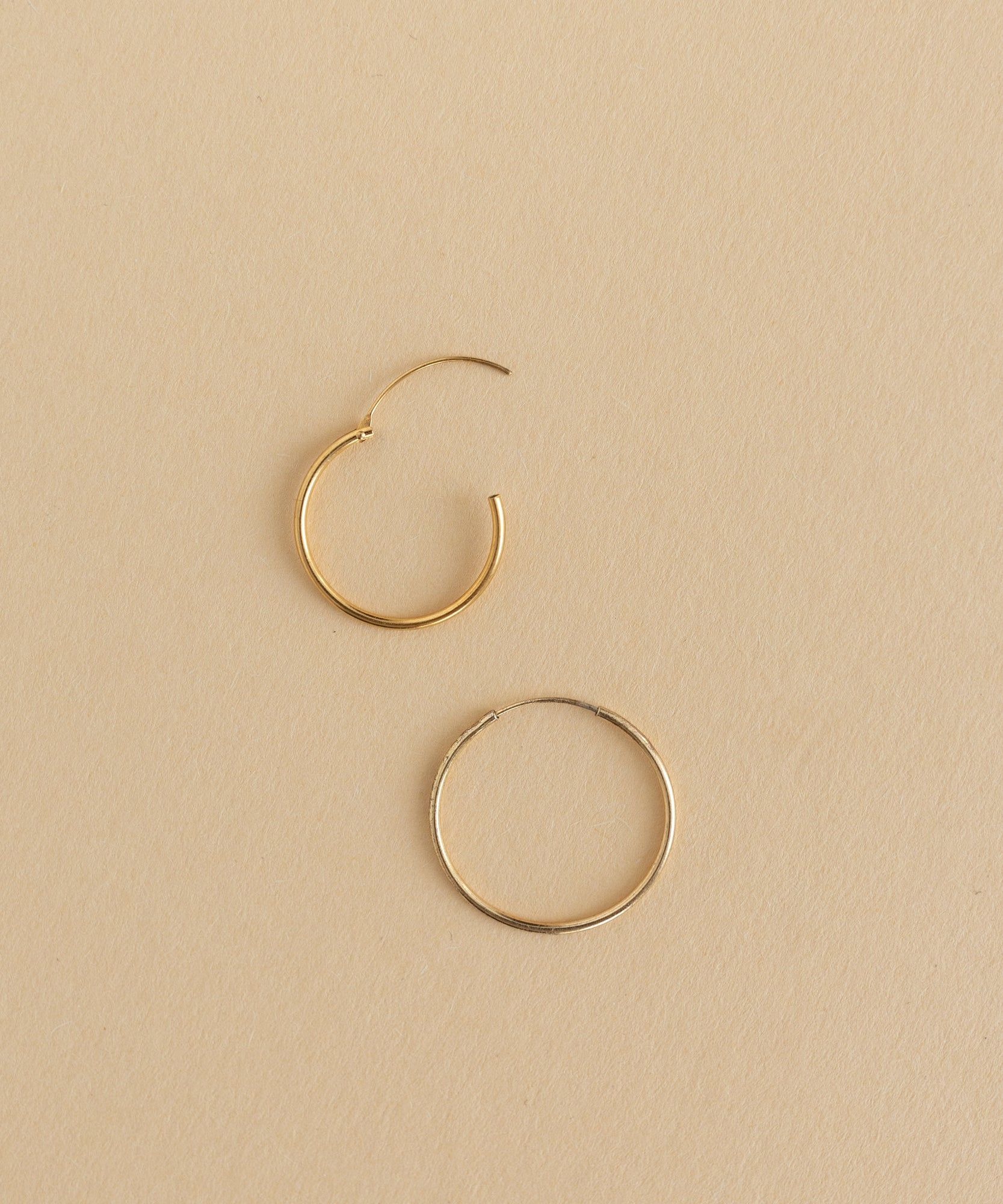 Two Gold Charm Earrings from WALD World on a beige surface.