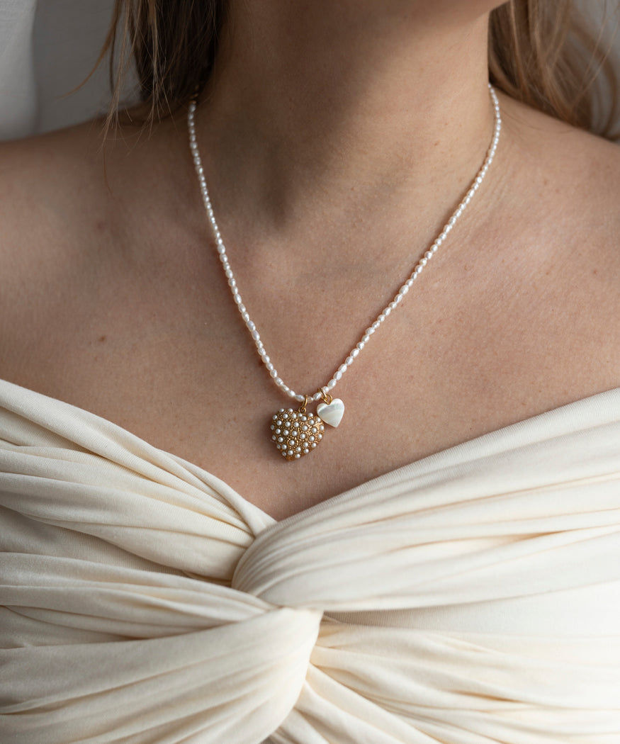 A woman wearing an off-shoulder cream dress and the WALD Berlin Rice Pearl I Am Love Double Heart Necklace featuring sweet water pearls, a gold-plated brass pendant, and a small heart charm.