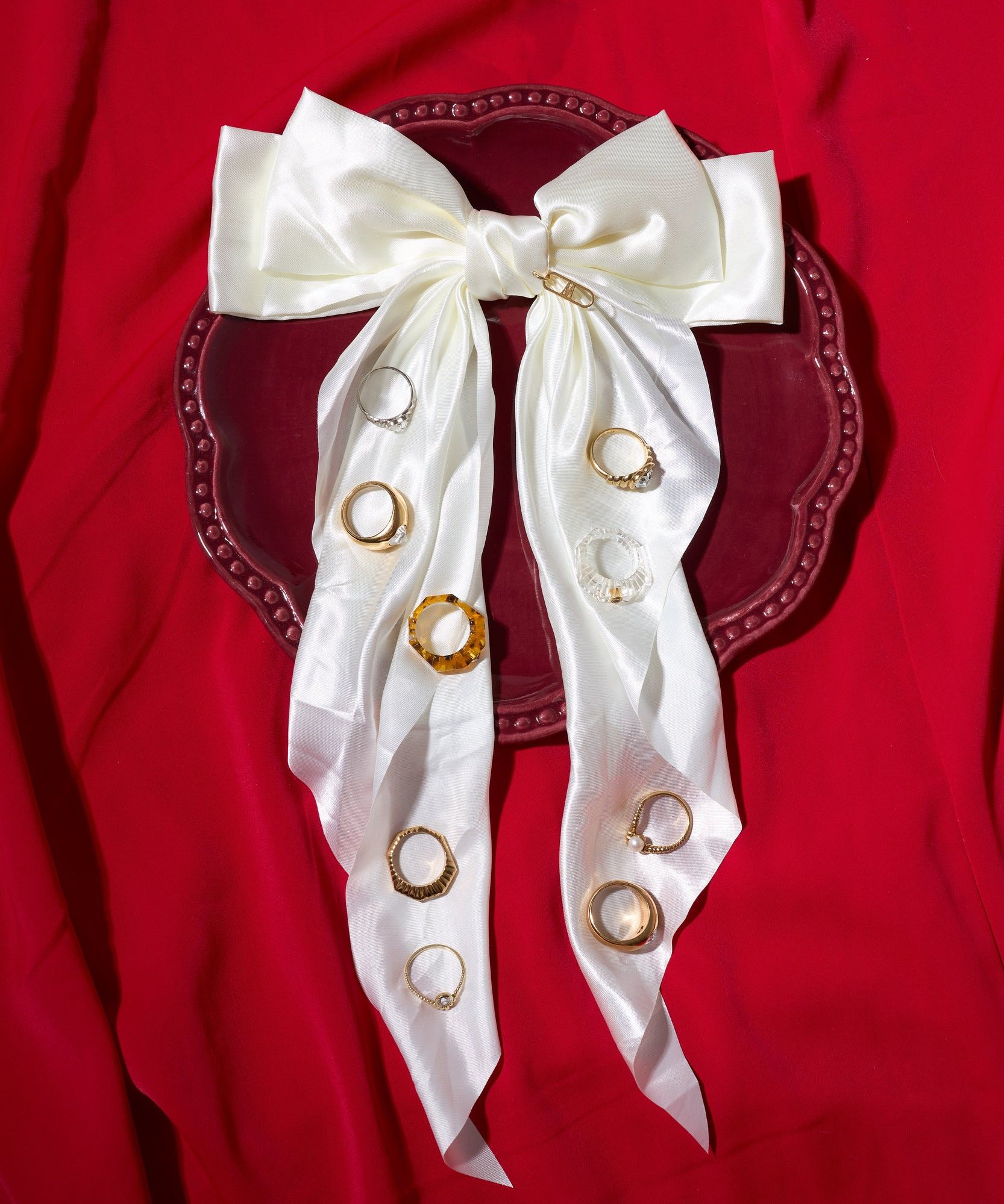 A large white satin bow is pinned to a red plate displaying nine assorted rings, including the WALD Berlin Pop Swarovski Ring, all set against a red fabric background.