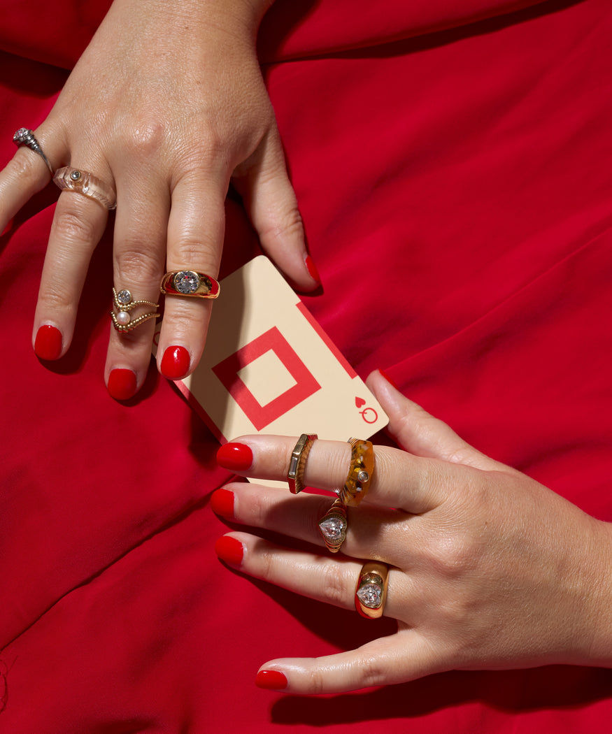 Two hands with red nail polish and multiple rings, including the WALD Berlin Mom Heart Ring in gold plated brass, hold a playing card with a red diamond against a red fabric background.