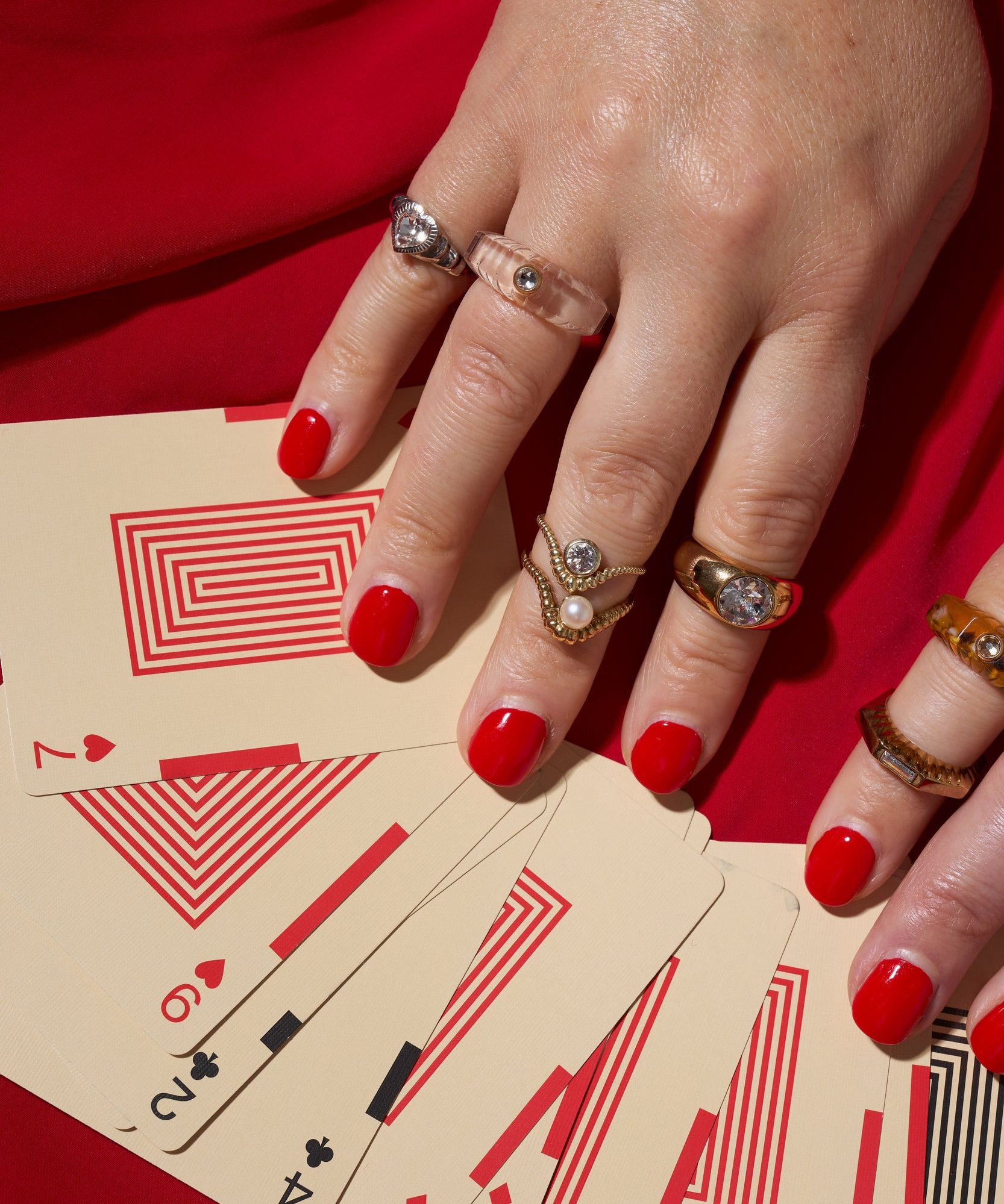A hand adorned with the WALD Berlin Shining Star Gold Ring rests on playing cards with geometric red patterns, spread out on a red surface; the ring features gold-plated sterling silver and Swarovski stones.