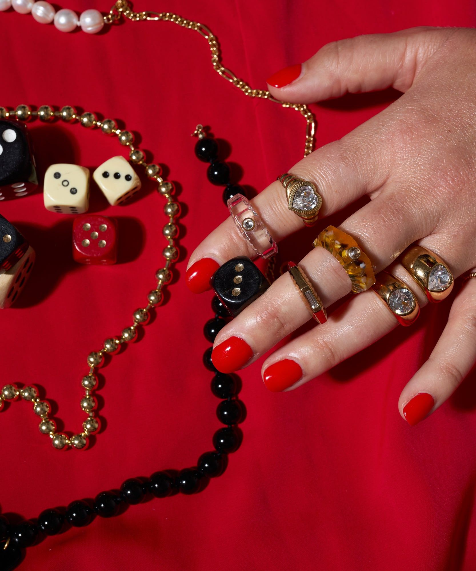 A hand with red nail polish displays the WALD Berlin Shining Star Gold Ring, stacked with other gold-plated sterling silver bands, resting on a red surface among beaded necklaces and assorted dice.