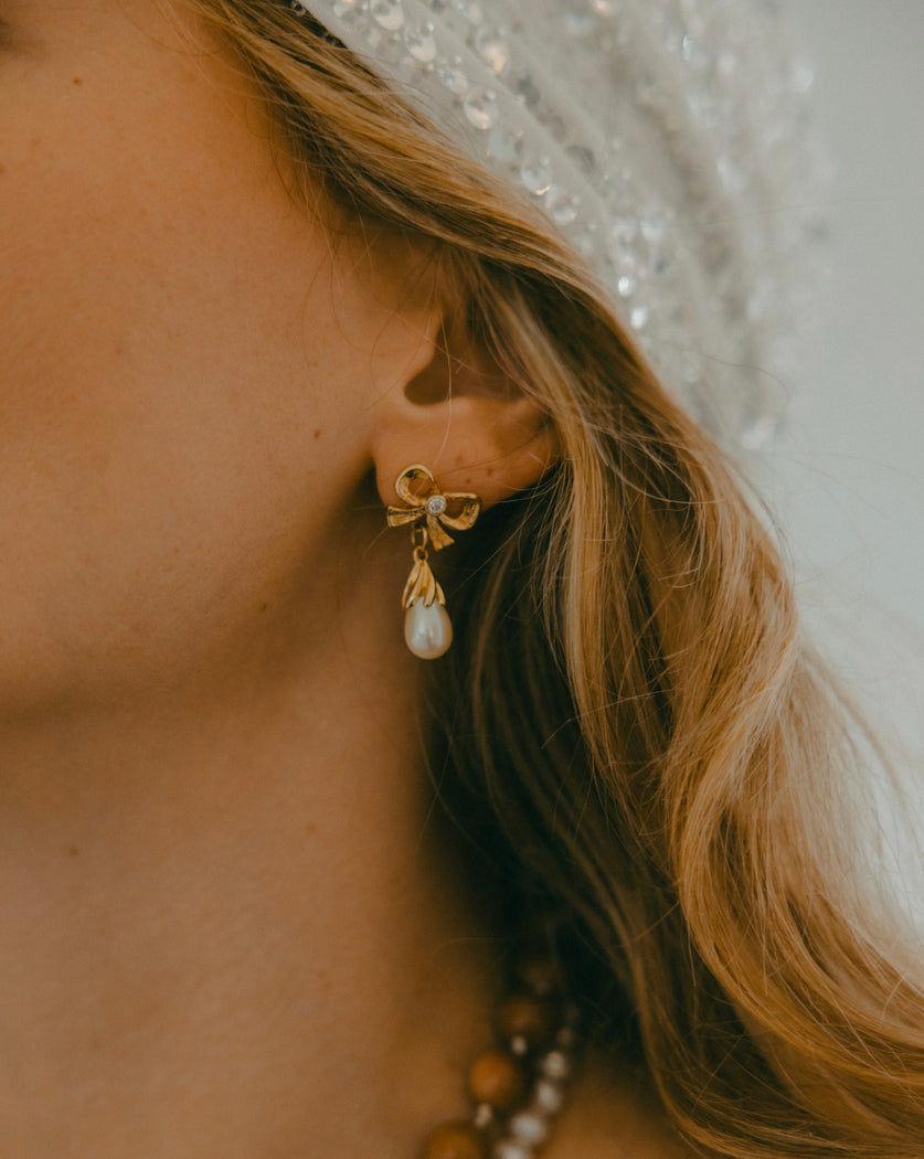 Close-up of a person wearing a sparkling headscarf and WALD Berlin's Happy Holidays Bow Earrings, featuring a pearl. Their hair is wavy and blond.