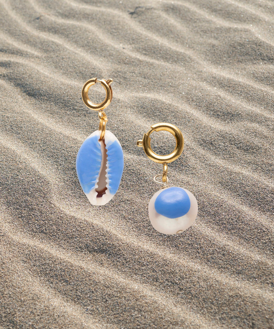 A pair of Beach Babe Charms earrings in uni-color blue and gold on the sand by WALD Berlin.