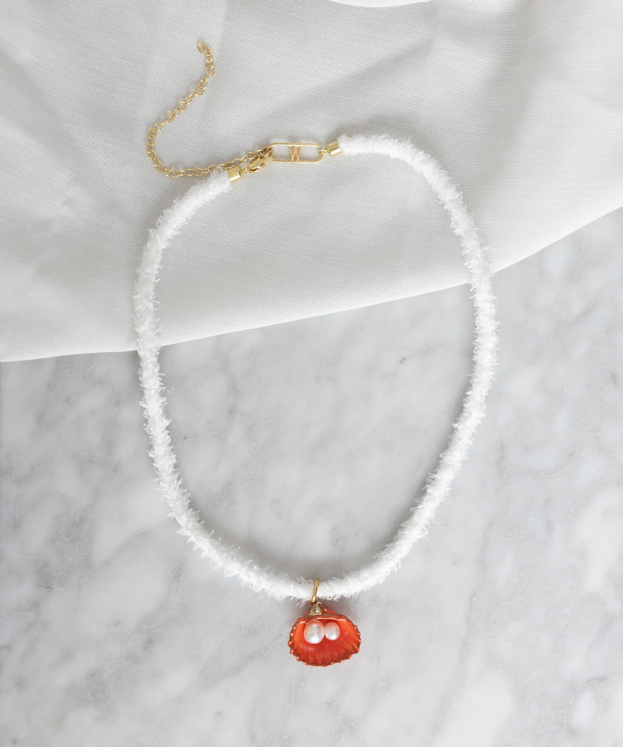 A white textured Fairtrade necklace with a gold clasp and chain, featuring a red seashell pendant and two pearls on a marble surface with a white fabric backdrop is the La Piscina Drop It Like Its Hot Necklace With Ribbon by WALD Berlin.