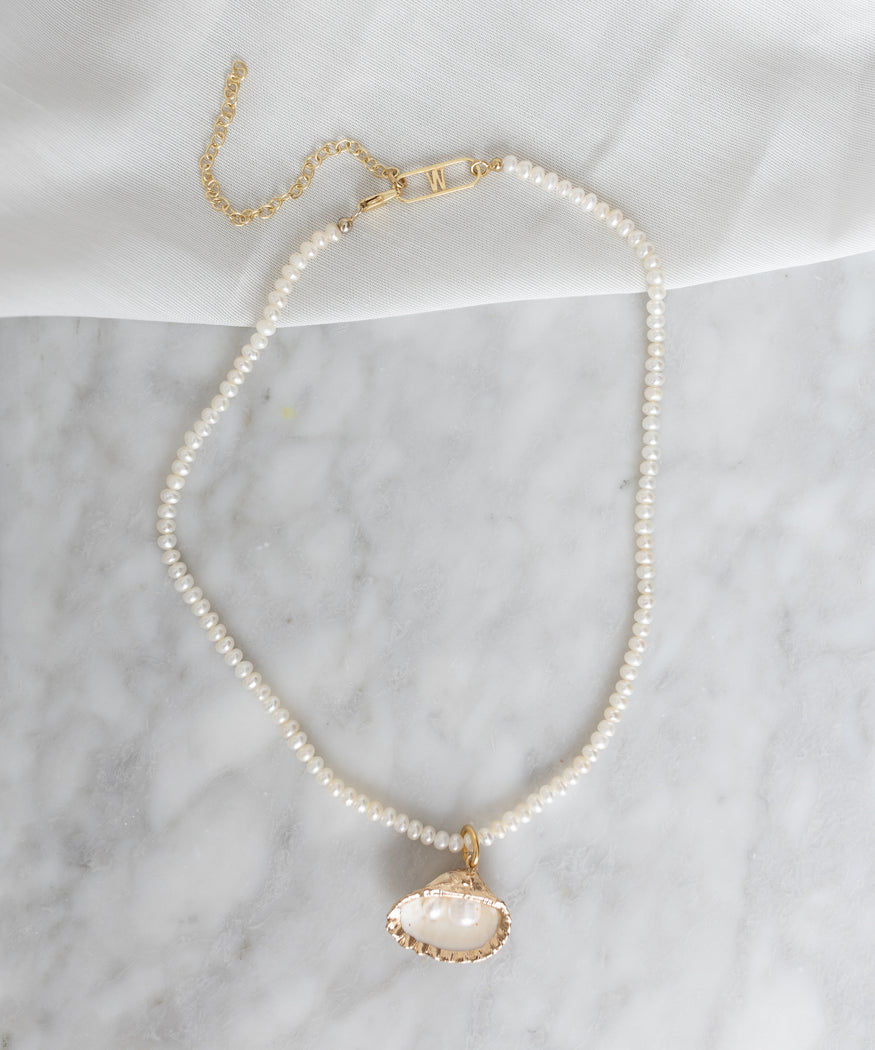 A WALD Berlin La Piscina Drop It Like Its Hot Pearl Necklace featuring lustrous pearls, a gold clasp, and a dangling gold-bordered white stone pendant is displayed on a white marble surface.