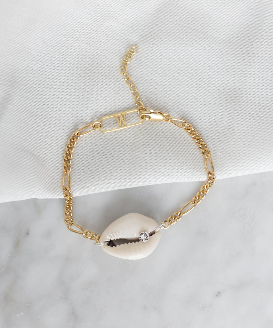 La Piscina Just a Friend Bracelet by WALD Berlin featuring a cowrie shell adorned with a small diamond accent, crafted from recycled gold and silver and placed on a white fabric background on a marble surface.