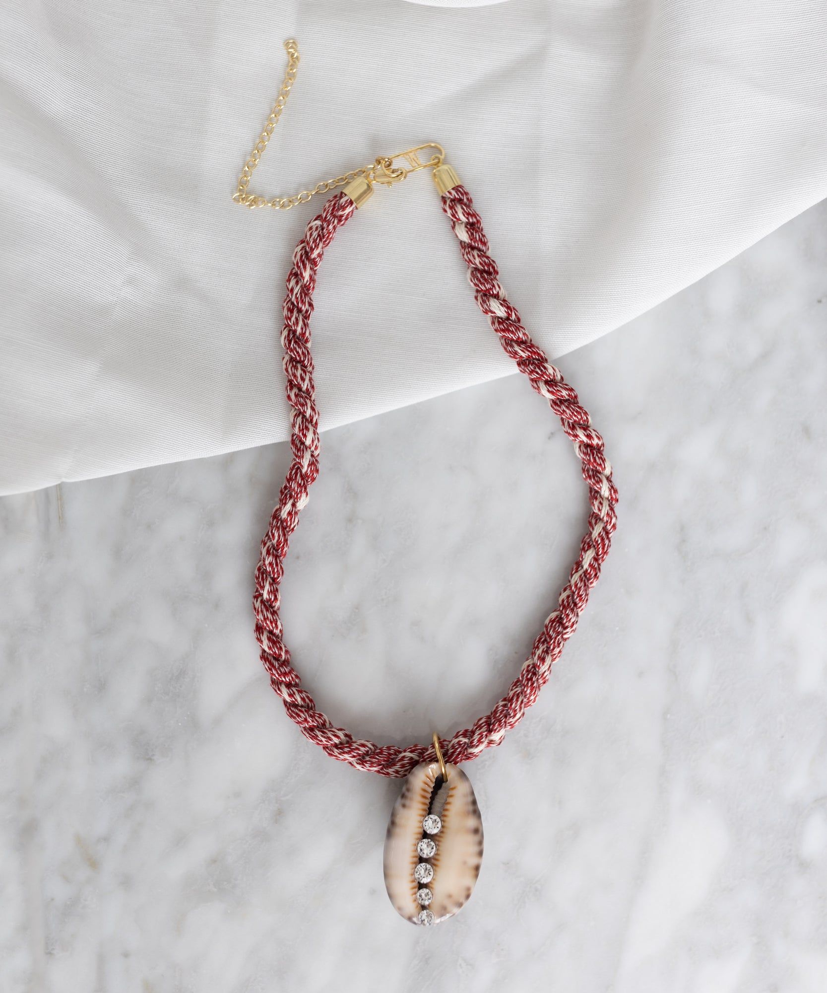 A red and white braided necklace with a seashell pendant featuring three embedded Swarovski stones, laid on a marble surface and partially draped over a white cloth, **WALD Berlin La Piscina Bootylicious Necklace With Ribbon**.