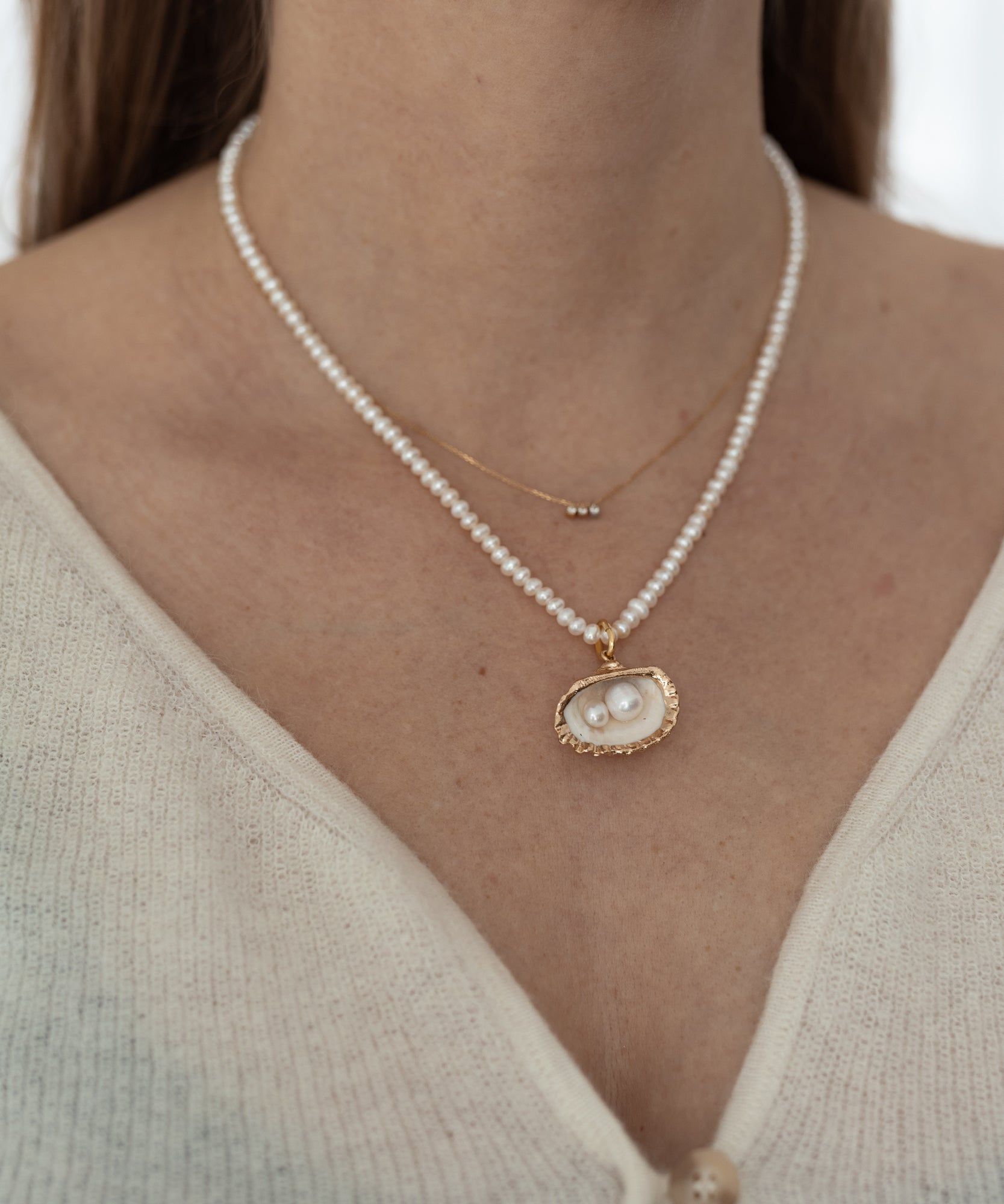 Close-up of a person wearing two Fairtrade necklaces: a small gold chain with a tiny pendant and a larger string of pearls with a gold-bordered pendant featuring two pearls. The person wears a light-colored top, including the La Piscina Drop It Like Its Hot Pearl Necklace by WALD Berlin.