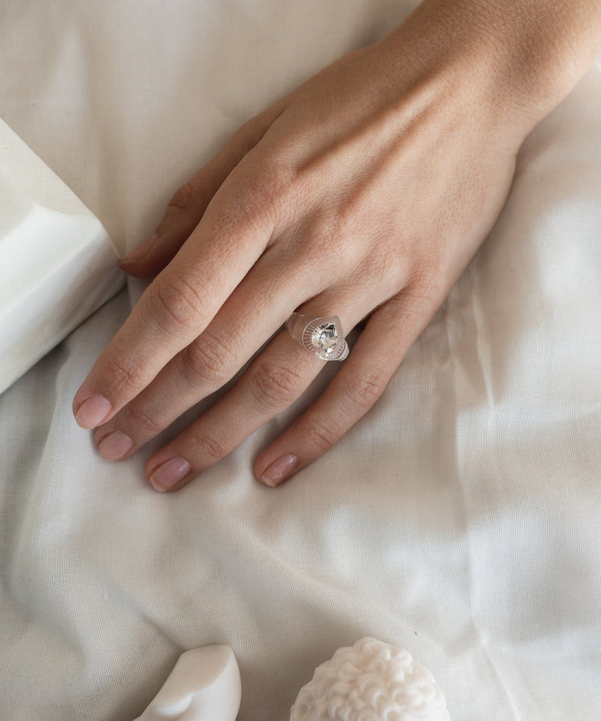 A woman's hand holding a Be My Lover Transparent Ring by WALD Berlin on a bed.