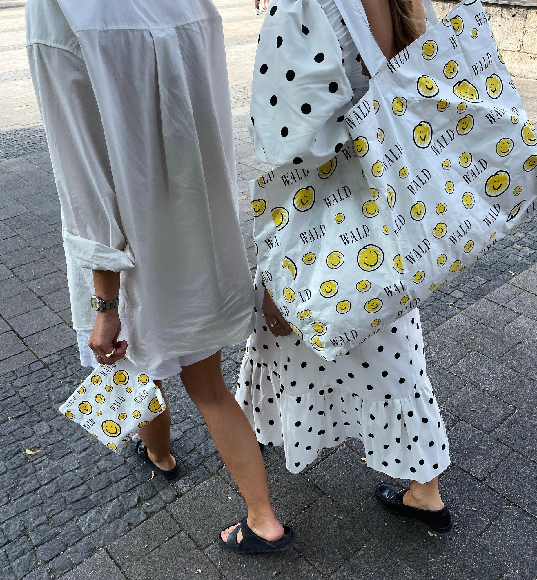 Two Berlin women flaunting jewelry while walking down the street with WALD Berlin Smilie Dude Shopper bags.
