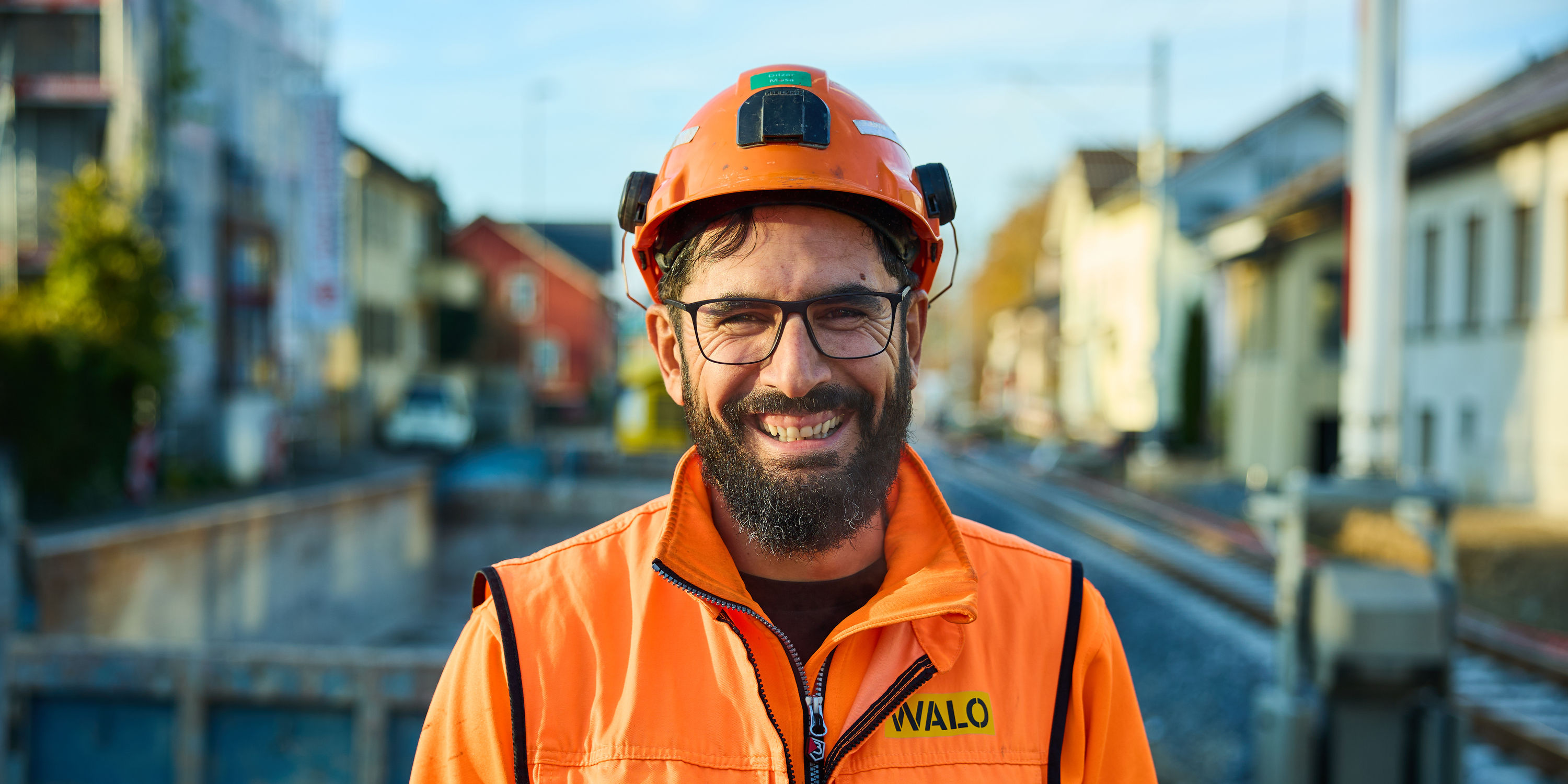 Un employé vêtu d'une tenue de sécurité orange et d'un casque sourit sur un chantier urbain.