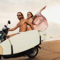 couple riding motorbike with surfboard on beach in bali, indonesia