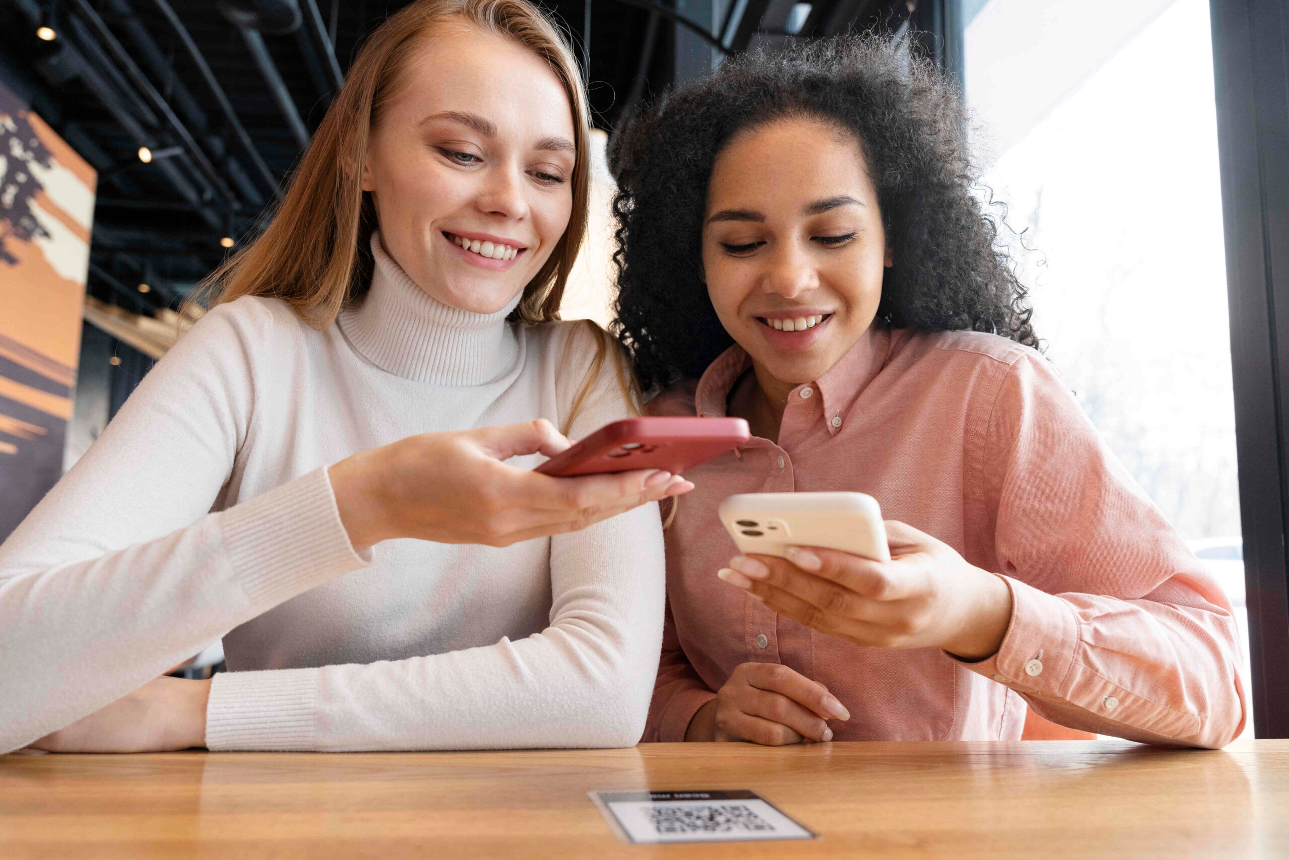 Duas jovens mulheres, a da esquerda é loira e de pele clara, já a da direita tem a pele escura e cabelo cacheado. As duas estão usando o Wi-Fi gratuito de um estabelecimento. Este recurso ajuda a captar clientes.