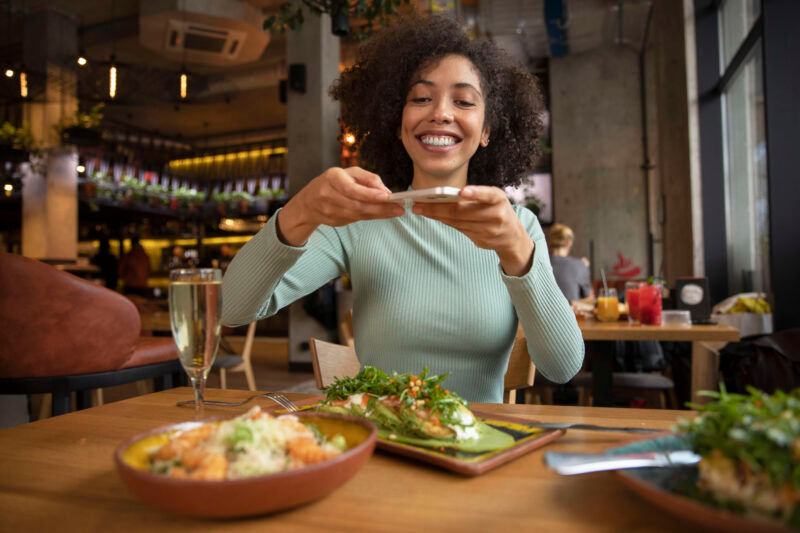Mulher sorrindo enquanto tira uma foto do prato de comida em um restaurante. Ela está sentada à mesa com duas refeições à sua frente, sorrindo para a câmera. A imagem destaca o ambiente moderno e confortável do restaurante, com o conceito de Automação para Restaurante com WiFi gratuito, proporcionando uma experiência prática e eficiente para os clientes.