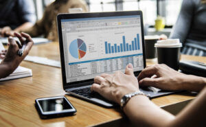 Pessoa utilizando um notebook com planilhas e gráficos de desempenho financeiro durante uma reunião com a equipe de vendas, em um ambiente de trabalho colaborativo.