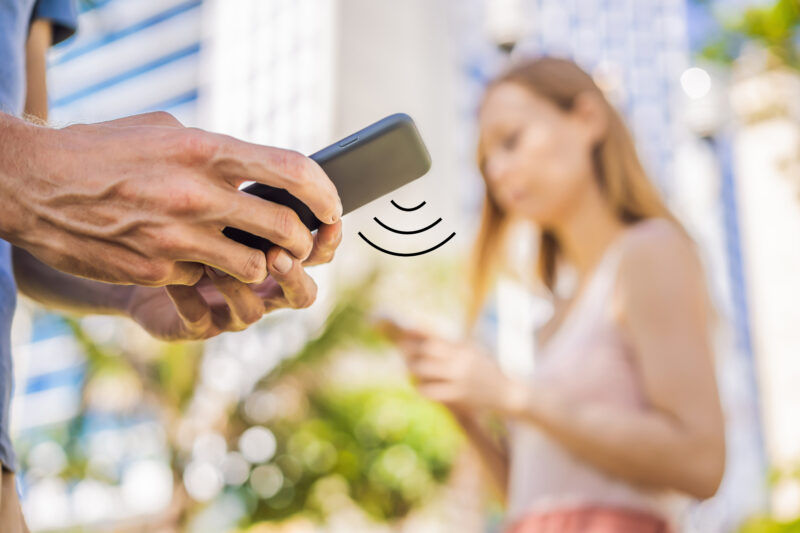 Dois usuários com smartphone em ambiente ao ar livre utilizam Wi-Fi público. A imagem destaca o gesto de um dos indivíduos interagindo com o telefone enquanto um símbolo de sinal de Wi-Fi aparece, sugerindo a utilização da tecnologia roaming WiFi.