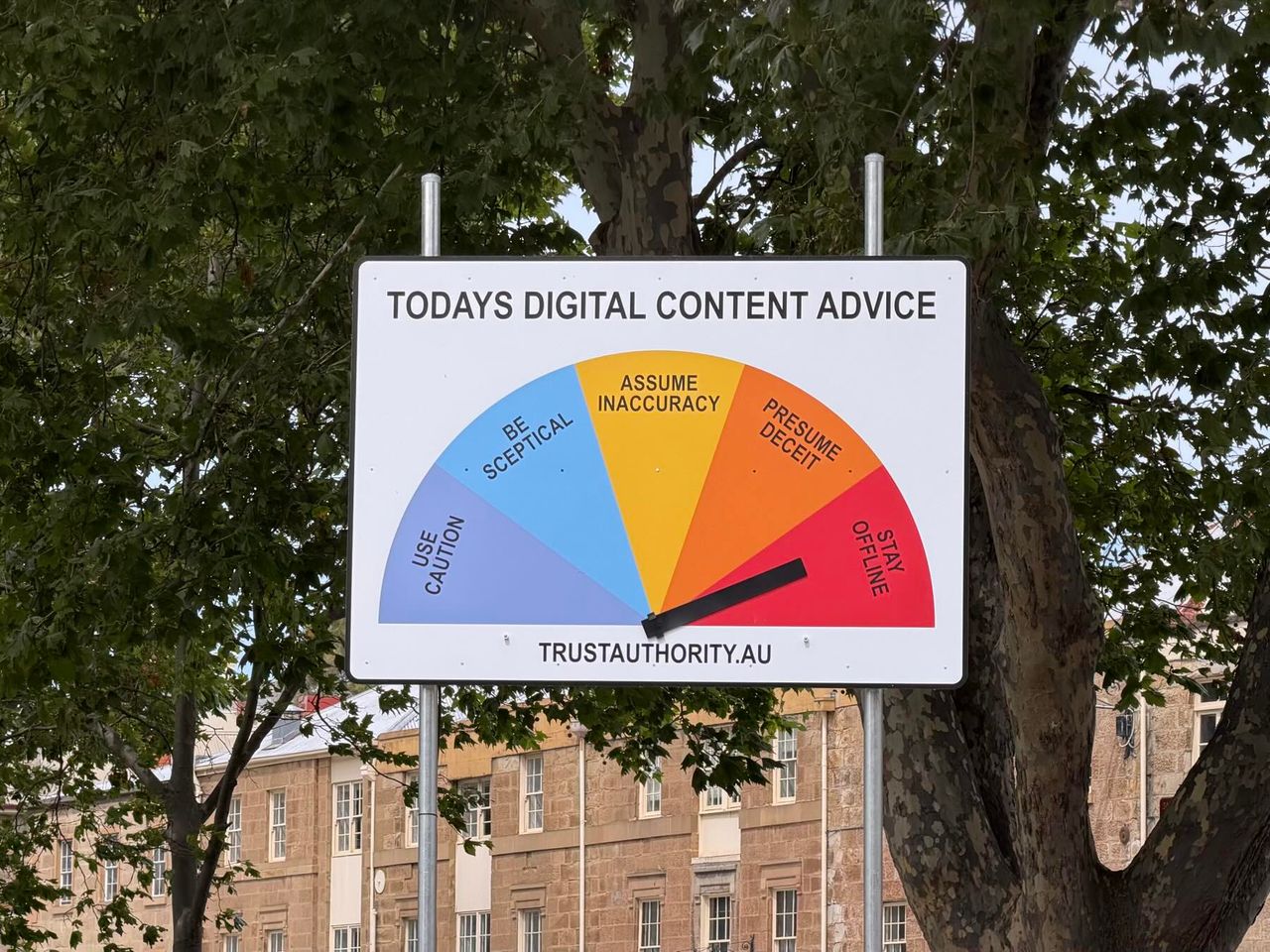 White sign with multi-coloured gauge and black needle under trees near a stone building.