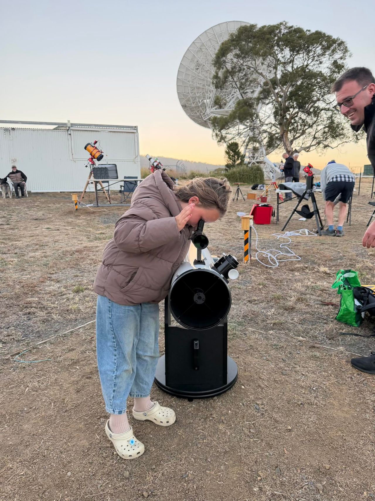 A woman in a brown jacket looking through a telescope in a field with other telescopes and people.