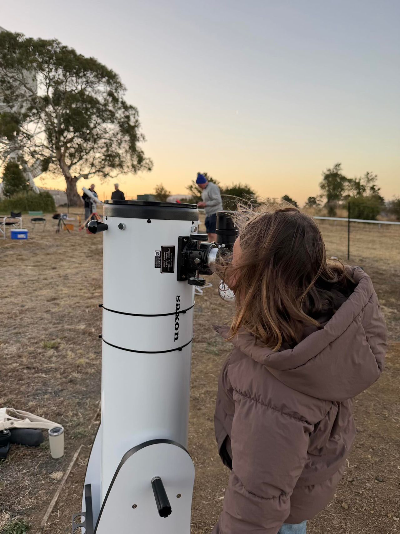 A woman in a brown coat looking through a telescope in a field at sunset.