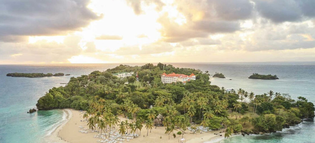 Vista aérea panorámica del lujoso resort Cayo Levantado en una isla tropical, mostrando la piscina frente al mar turquesa, playas de arena blanca bordeadas de palmeras y pequeños islotes rocosos en Samaná, República Dominicana.