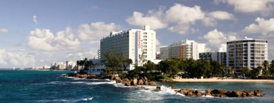 Scenic coastline view of the Condado Plaza Hotel beachfront with blue ocean water, rocks, and a cloudy sky in San Juan, Puerto Rico.