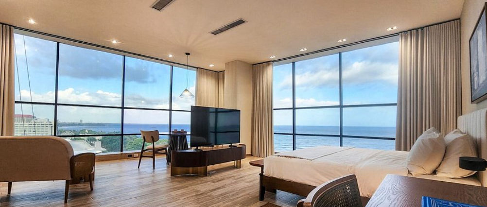 Alt text: Modern and spacious room at Mauad Hotel Boutique featuring large floor-to-ceiling windows, a double bed, sitting area, and a panoramic ocean view of Santo Domingo.