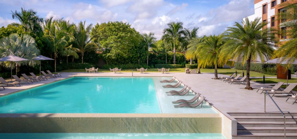 Modern facade of the AC Hotel by Marriott Punta Cana featuring the swimming pool, lounge chairs, and palm trees on a sunny day