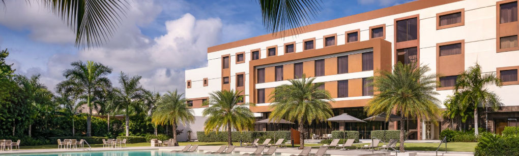 Modern facade of the AC Hotel by Marriott Punta Cana featuring the swimming pool, lounge chairs, and palm trees on a sunny day