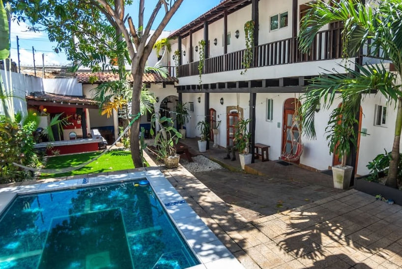 Patio interior colonial con piscina pequeña, hamaca, árboles y balcones de madera en un hostal de Santo Domingo.