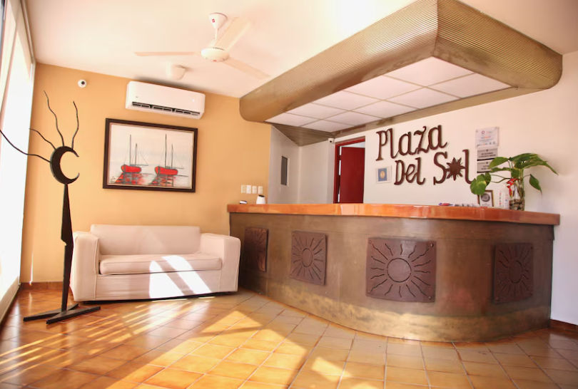 Reception desk at Aparta Hotel Plaza del Sol featuring a wooden counter, a white sofa, air conditioning, and the hotel name on the back wall.