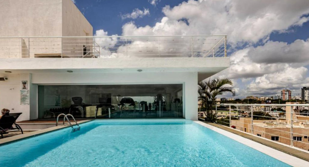 Terraza en la azotea con piscina, tumbonas y mesas con vistas al mar bajo un cielo azul despejado / Rooftop terrace featuring a swimming pool, lounge chairs, and dining tables with ocean views under a clear blue sky