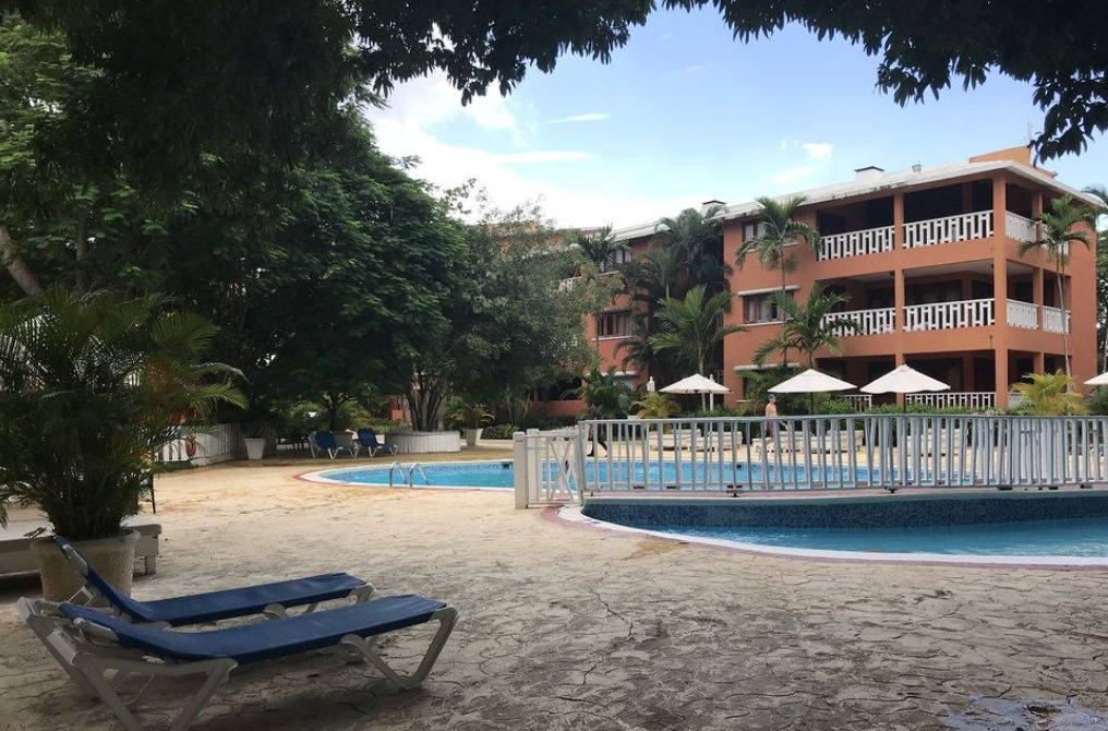 Piscina al aire libre rodeada de palmeras tropicales, bar junto a la piscina y edificios de habitaciones en el hotel Bellevue Dominican Bay / Outdoor swimming pool surrounded by tropical palm trees, a poolside bar, and guest room buildings at Bellevue Dominican Bay hotel