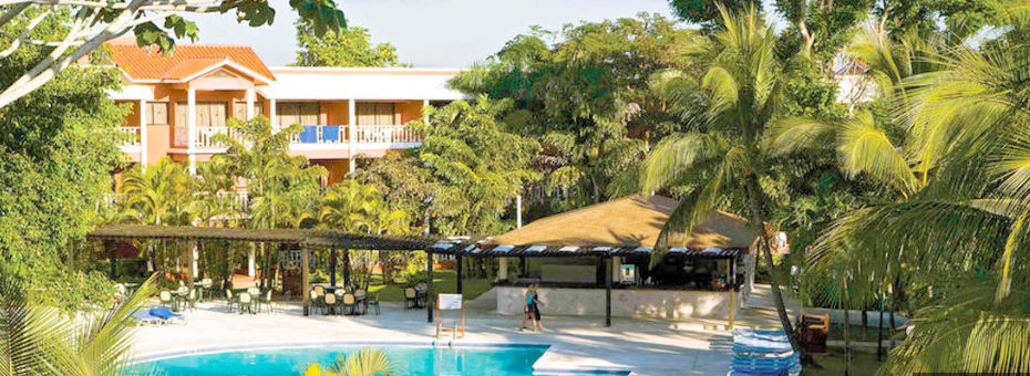 Outdoor swimming pool surrounded by tropical palm trees, a poolside bar, and guest room buildings at Bellevue Dominican Bay hotel