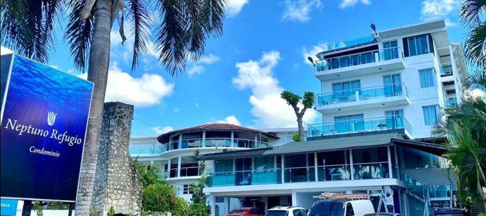 Blue outdoor sign reading "Neptuno Refugio Condominio" in front of a modern white multi-story residential building with glass balconies under a partly cloudy sky.