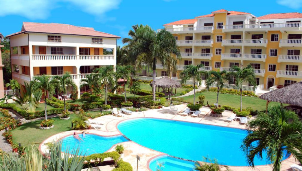 Aerial view of the swimming pool and yellow residential buildings at Residencial Las Palmeras de Willy in Boca Chica, surrounded by tropical gardens and palm trees under a blue sky.
