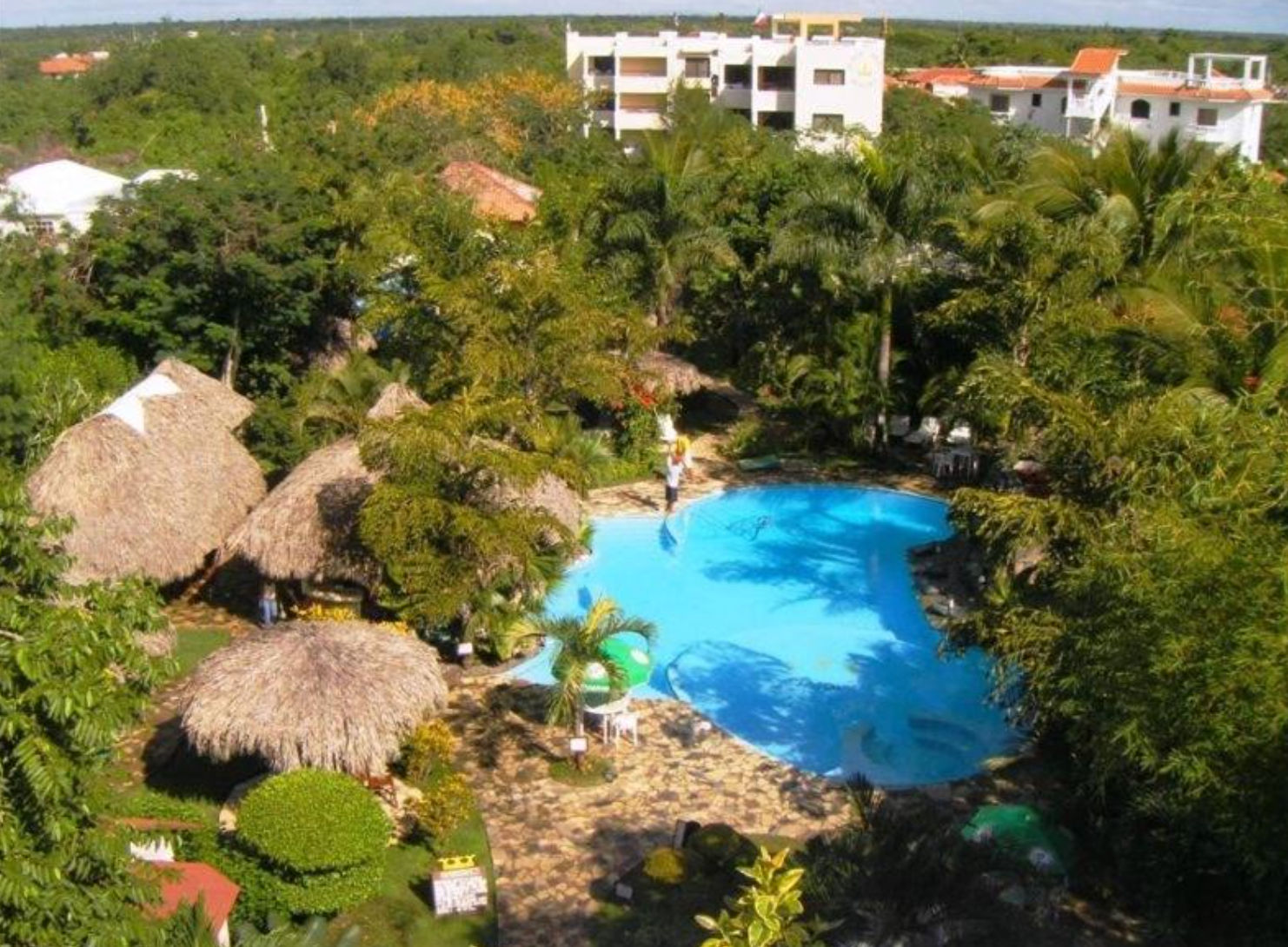 Aerial view of the swimming pool at Plaza Real Resort Juan Dolio, surrounded by tropical palm trees, thatched umbrellas, and hotel buildings.