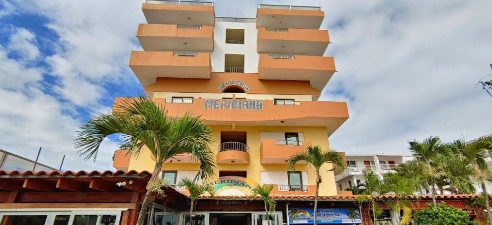 Facade of the Meridian Residence Juan Dolio hotel with orange balconies and palm trees under a partly cloudy sky in the Dominican Republic.