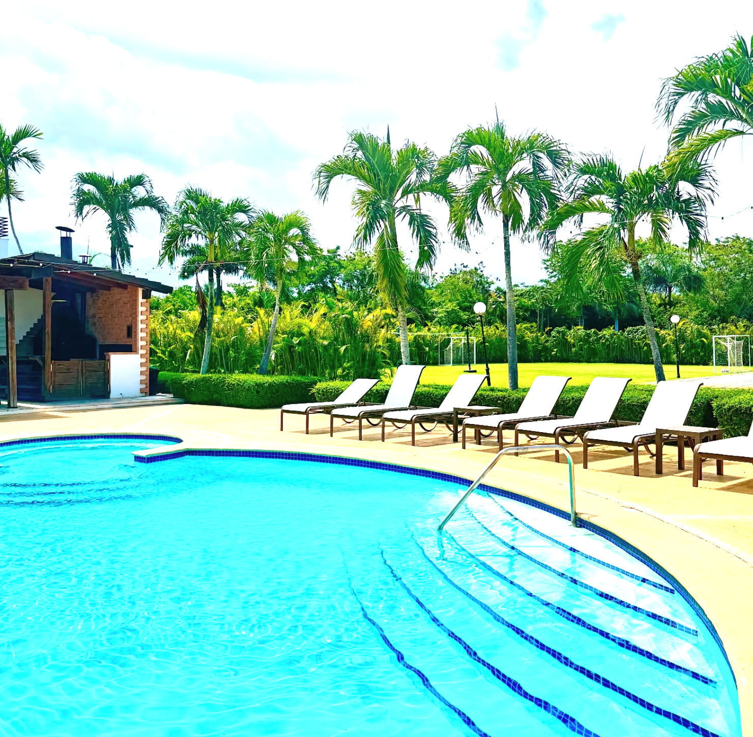 Área de piscina y restaurante Cohoba en Sybaris Suites & Residences con un puente peatonal de madera, palmeras y vegetación tropical bajo un cielo nublado