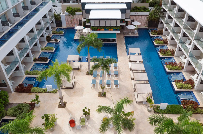 presenta una vista aérea del patio interior de un lujoso complejo turístico, que cuenta con una destacada piscina azul en forma de T. La piscina está bordeada por edificios modernos de varios pisos con balcones. Está bien equipado con numerosas tumbonas a