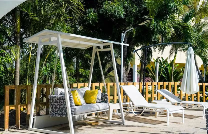Outdoor dining area of Restaurant Soficu Eden featuring tables and chairs under a roof and wooden pergola, located next to a blue swimming pool with a dolphin statue, a palm tree, and a white hotel building in the background.