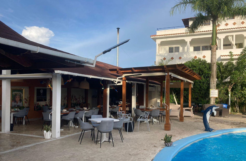Outdoor dining area of Restaurant Soficu Eden featuring tables and chairs under a roof and wooden pergola, located next to a blue swimming pool with a dolphin statue, a palm tree, and a white hotel building in the background.