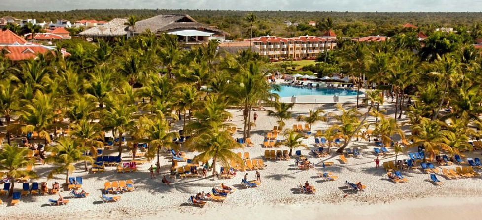 Vista elevada de un hotel con palmeras, playa, piscina y un paisaje exuberante, con tumbonas de playa en la arena.