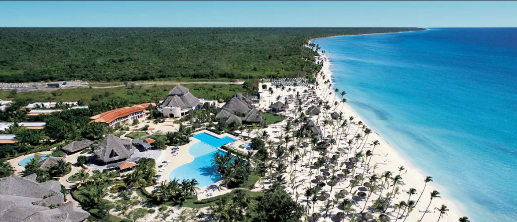 Aerial photograph of a large beachfront resort with prominent blue swimming pools, red-roofed buildings, and numerous large thatched-roof palapa structures. The complex is set between a vast tropical forest and a white sand beach with palm trees and clear
