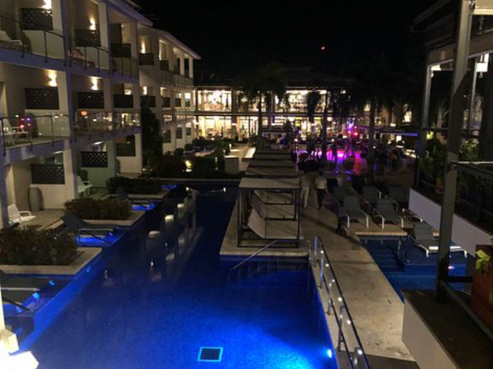 Night view of a resort swimming pool illuminated in blue with multi-level guest rooms, lounge chairs, and palm trees in the background.